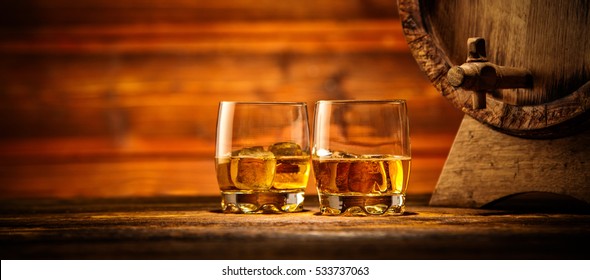 Two glasses of whiskey with ice cubes served on wooden planks with keg. Vintage countertop with highlight and a glass of hard liquor