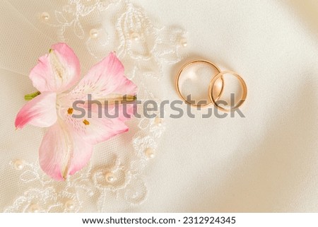 Two gold engagement rings and pink astromeria flower on a beige satin background. Wedding background. A copy space