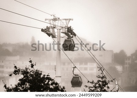 Two gondolas of the Vilanova de Gaia cable car suspended on hanging steel cables descending and ascending under a cloudy white sky with the classic neighborhood of Gaia shrouded in fog. Monochrome.