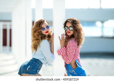 Two gorgeous brunette girlfriend are walking along the embankment, having fun, smiling, laughing. With makeup and curls. Red lipstick and sunglasses. Summer handbags and denim shorts.