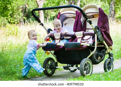 Two Gorgeous Girls Twins Walking On Green Grass With Pleasure On A Double Buggy