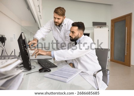 Two GP professionals in white lab coats collaborating at computer, analyzing digital medical information, patient data or diagnostic results, engaged in medical case consultation and x-ray scan review