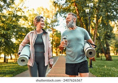 Two gray-haired seniors dressed in sportswear go to exercise outdoors in the park, carry exercise mats, look at each other and talk.Modern active beautiful cheerful sincere carefree pensioners. - Powered by Shutterstock