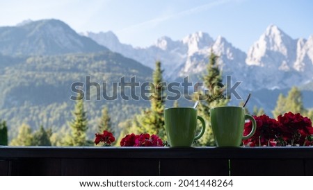 Two green coffee cups sitting on  a wooden balcony fence with beautiful view of mountains and pine trees on a sunny day.