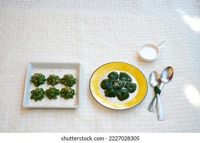 Two green Thai sweets are in coconut milk, topping with sesame seeds.  A pair of small spoon is by the side of ceramic plate.  White textile table cloth as background. - Shutterstock ID 2227028305