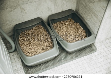 Two grey litter boxes filled with wood pellet litter, placed on a tiled floor. A textured mat borders the boxes, and a grey corrugated hose is visible to the left.Clean, functional pet accessory setup