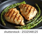 Two grilled chicken breasts on a grill pan, with asparagus spears in the background