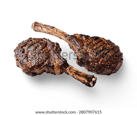 Two grilled tomahawk ribeye steaks seasoned with herbs and pepper, isolated on a plain white background.