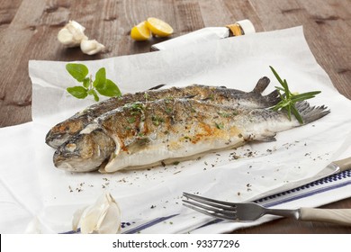 Two Grilled Trouts With Lemon Pieces, Fresh Herbs And Garlic On White Baking Paper On Kitchen Towel On Wooden Table.