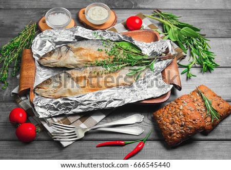 Two grilled whole fish trout on an aluminum foil on a gray wooden background. Additional ingredients for grilled fish greens rosemary, thyme, sage, red pepper, bread, fish sauce tartar. 