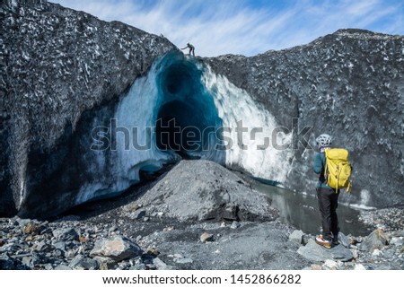 Two guides looking at a massive ice cave on the Matanuska Glacier in Alaska. Before entering the cave, one guide on top knocks down rocks that may fall from above.