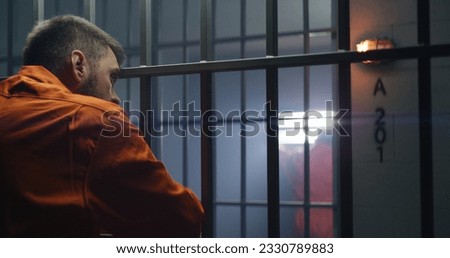 Two guilty men serve imprisonment term together in prison. Elderly criminal stands in jail cell, holds hands on bars and talks with neighbour. Offenders in detention center or correctional facility.