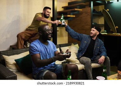 Two Guys Clinking With Bottles Of Beer While Their African American Buddy With Joystick Sitting On Couch And Playing Video Game