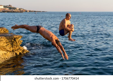 Two Guys Diving From A Cliff. Muscular Guys, Bomb Jump And Swimming Trunks