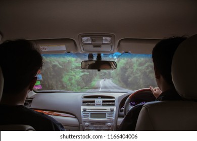 Two Guys Inside The Car Driving On The Country Roadway Between Fields With Green Grass And Tree Mountains On The Cloudy Sky Background In Thailand.