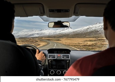 Two Guys Inside The Car Driving On The Country Roadway Between Fields With Brown Grass And Snowy Mountains On The Cloudy Sky Background In Iceland. Sun Is Shining. Shoot From The Back. Horizontal.
