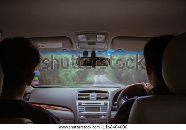 Two guys inside the car driving on the country\
roadway between fields with green grass and tree mountains on the\
cloudy sky background in\
thailand.
