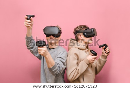 Two guys play VR games in virtual reality helmets on pink background, hold controllers and look away. Two friends in VR helmets play video games. Isolated. VR gaming with friends.