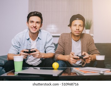 Two Guys Playing On The Console Sitting On The Couch. European And African Friends Had A Party