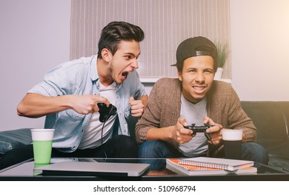 Two Guys Playing On The Console Sitting On The Couch. European And African Friends Had A Party. Victory Celebration