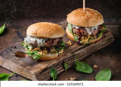 Two Hamburgers With Beef Burger Cutlet, Fried Onion, Spinach, Ketchup Sauce And Blue Cheese In Traditional Buns, Served On Wood Chopping Board Over Dark Wooden Background.