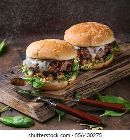 Two Hamburgers With Beef Burger Cutlet, Fried Onion, Spinach, Ketchup Sauce And Blue Cheese In Traditional Buns, Served On Wood Chopping Board With Knife And Fork Over Dark Background. Square Image