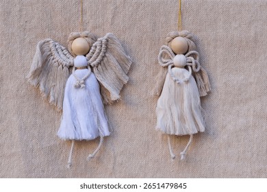 Two handmade macrame angels in neutral tones, featuring wooden heads and textured cords. Hanging against natural fabric background. Ideal for rustic, Christmas decor, DIY crafts, and natural themes