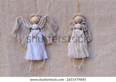 Two handmade macrame angels in neutral tones, featuring wooden heads and textured cords. Hanging against natural fabric background. Ideal for rustic, Christmas decor, DIY crafts, and natural themes