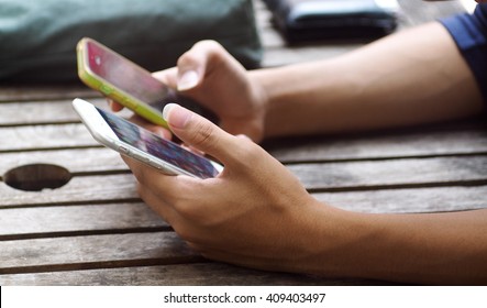 Two Hands Of An Asian Boy Holding Two Mobile Phones On A Wooden Table Trying To Connect Or File Transfer Between The Both Mobile Handy Smart Phones That Reflect Green Environment On The Touch Screens