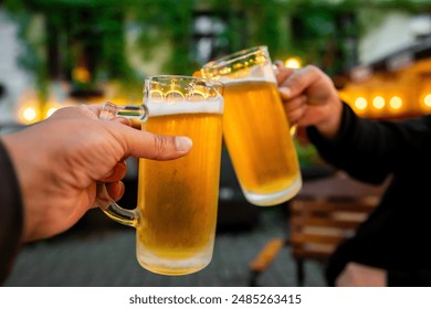 Two hands clinking beer mugs in a toast, with a blurred background of lights and greenery - Powered by Shutterstock