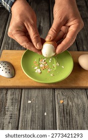 Two hands are cracking an egg on a green plate. The shell breaks, and small pieces fall onto the plate. An uncracked egg is nearby on a wooden surface.