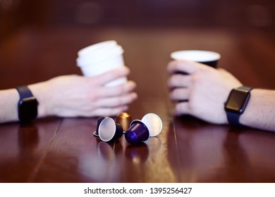 Two hands with equal black electronic wrist watches, holding cups of coffee, white and black, on the wooden table with replaceable cartridges with coffee on front. Date or friends meeting.Copy space.