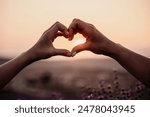 Two hands forming a heart shape in front of a purple field. The sun is setting in the background, creating a warm and romantic atmosphere.