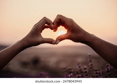 Two hands forming a heart shape in front of a purple field. The sun is setting in the background, creating a warm and romantic atmosphere. - Powered by Shutterstock