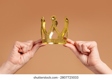 Two hands gently hold a small gold crown against a warm neutral background, evoking themes of leadership, achievement, celebration, royalty, recognition and personal triumph.