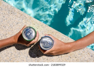 Two Hands Hold Cans Of Cold Drink In The Pool On A Hot Day. The Concept Of Vacation In A Friendly Company. Top View, Selective Focus On The Bank.