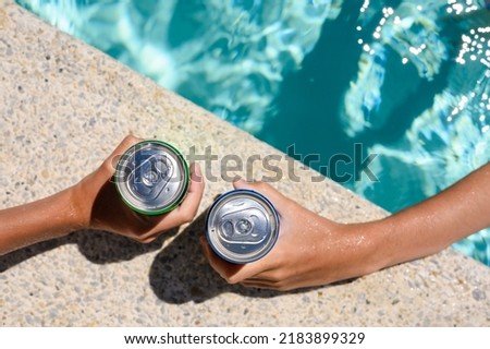 Two hands hold cans of cold drink in the pool on a hot day. The concept of vacation in a friendly company. Top view, selective focus on the bank.