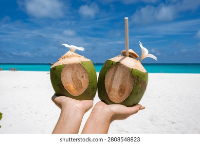 Two hands hold Coco Palm Dhuni Kolhu Maldives coconuts with frangipani and a straw on a white sand beach by a turquoise lagoon in bright midday sun, symmetrical view.