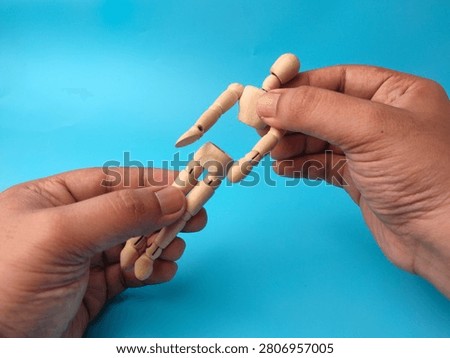 Two hands hold the separate torso and leg sections of a broken wooden artist mannequin on blue background.