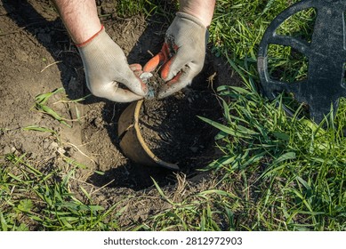Two hands hold a small object while digging in the ground with metal detector nearby. The location has grass and soil with a potential treasure buried underground.