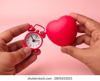 Two hands hold a small red mechanical alarm clock next to a matching red stress ball heart over pink.