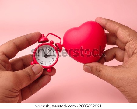 Two hands hold a small red mechanical alarm clock next to a matching red stress ball heart over pink.