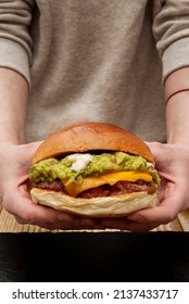 Two Hands Holding A Beef Burger With Cheddar Cheese And A Lot Of Mexican Guacamole On A Wooden Table And Black Slate