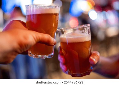 Two hands holding glasses of amber-colored beer with frothy heads, set against a blurred background suggesting a lively bar or pub atmosphere. Perfect for themes of socializing and nightlife. - Powered by Shutterstock