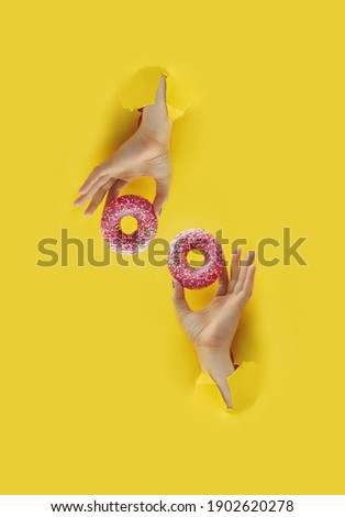 two hands holds delicious pink round glazed donut with sprinkle in torn yellow bright paper hole, vertical popular food blog content, selective focus