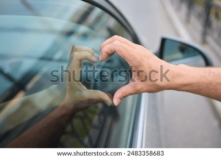 Two hands making a heart shape on opposite sides of a car window represent love and connection between individuals