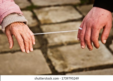 Two Hands Old And Young At An Elongated White Thread. Close-up White Thread Connecting Hands Together. Grandmother's Hand Tied To The Hand Of Her Grandson. Conceptual Photo.