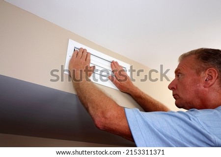Two hands on a closed air vent outflow to reduce air circulation and flow out of a home furnace air duct. Adult male has two hands on closed home furnace white rectangle wall air vent duct on a wall