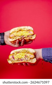 Two hands presenting a large sandwich burger sliced in half, revealing layers of ham, melted cheddar, pickles, and coleslaw against a bright red background, emphasizing indulgent textures 