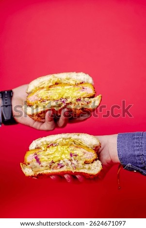 Two hands presenting a large sandwich burger sliced in half, revealing layers of ham, melted cheddar, pickles, and coleslaw against a bright red background, emphasizing indulgent textures 